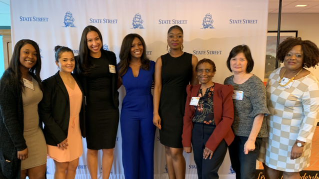 8 smiling women standing in front of a step and repeat banner
