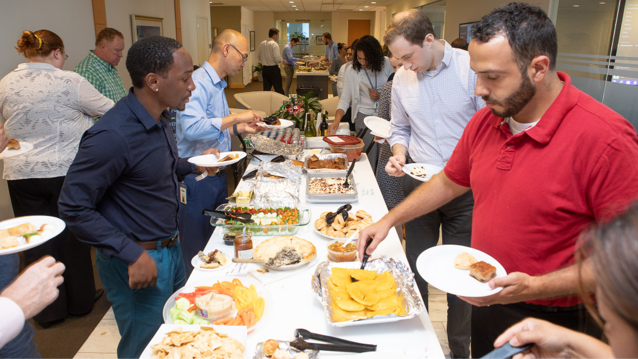 four male employees standing at a table filled with food as they fill their plates during the Inclusion Council international potluck
