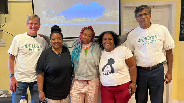 Two male employees wearing FHLBank Boston t-shirts standing next to three smiling women with a large screen behind them.