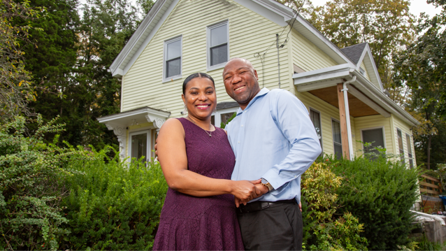 Woman and man standing next to each other outdoors in front of their home while holding hands and smiling.