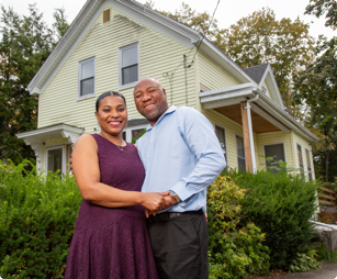 Woman and man standing next to each other outdoors in front of their home while holding hands and smiling.