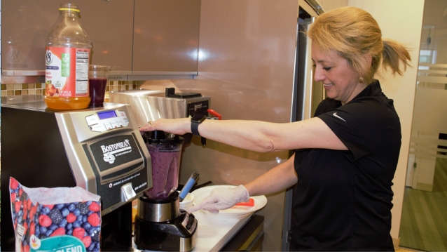 a smiling woman wearing a black shirt standing at a blender and blending a purple smoothie