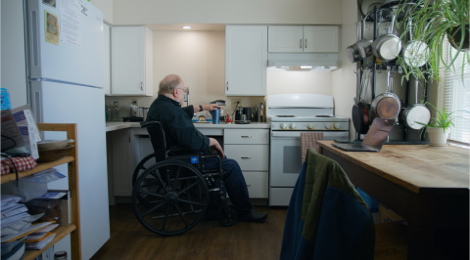 Man in wheelchair by the sink area of a kitchen with his hand placed firmly on the top of on insulated coffee mug. A laptop sits on a table to his right with pots and pans hanging on a rack near it.