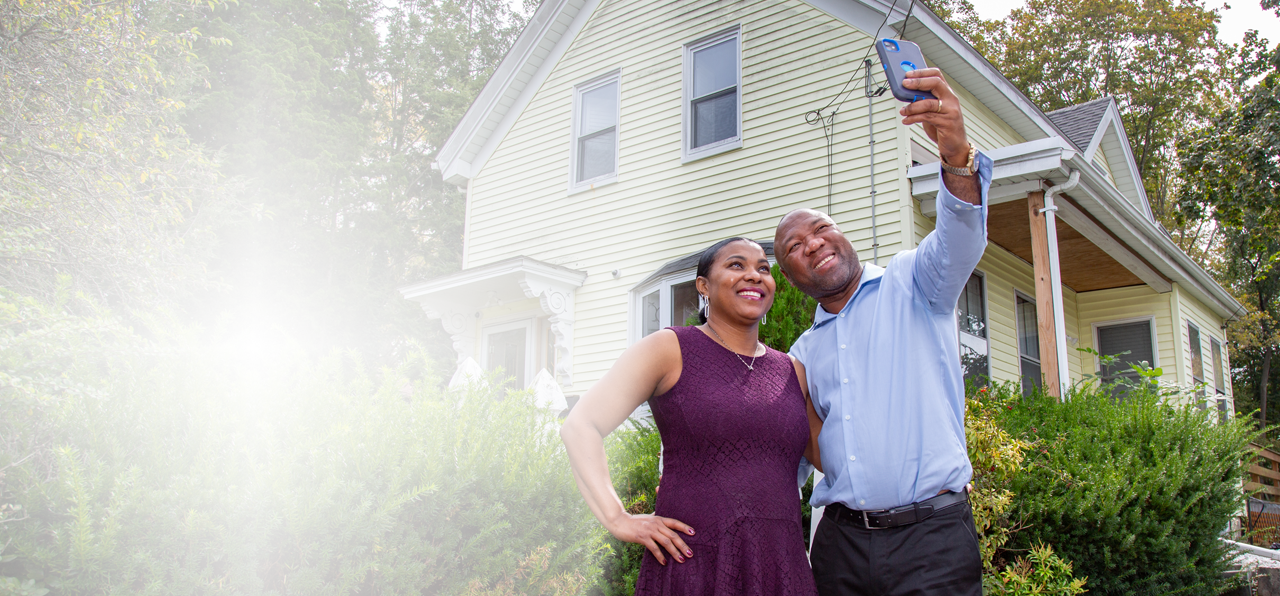 Man and woman standing next to each other outdoors in front of their home while taking a selfie