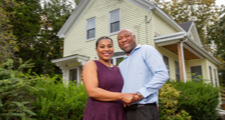 Woman and man standing next to each other outdoors in front of their home while holding hands and smiling.