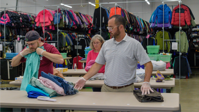 two FHLBank Boston male employees standing at a table with folded clothing as one of them folds a shirt