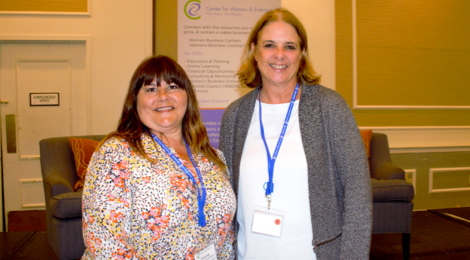 two female FHLBank Boston employees standing side by side smiling in front of a banner at a women's conference