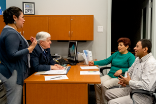 female standing near desk with man seated at desk and male and female couple seated in front of desk looking at seated male