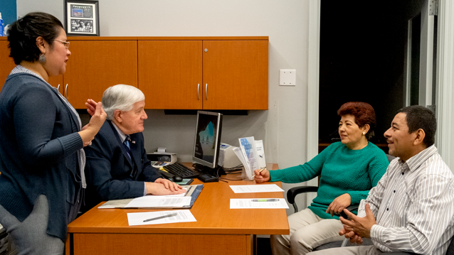 female standing near desk with man seated at desk and male and female couple seated in front of desk looking at seated male