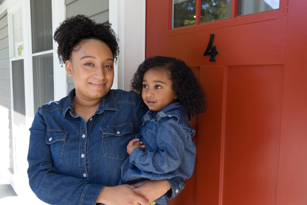 mother holding her young daughter as she stands at the front door of her home