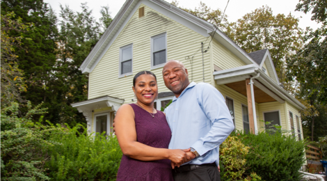 Woman and man standing next to each other outdoors in front of their home while holding hands and smiling.