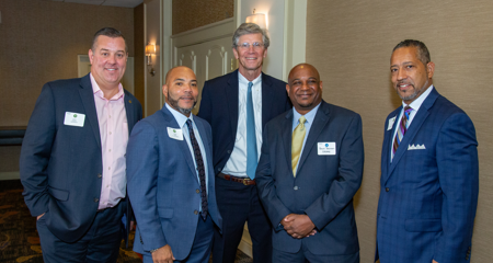 Five smiling men in business attire standing next to each other in the lobby area outside of a hotel ballroom.