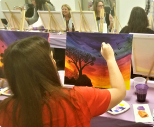 female employee seated while she is painting a colorful scene on a canvas